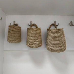 May include: Three brown crocheted hanging baskets with rope handles. The baskets are hanging from hooks on a white wall.