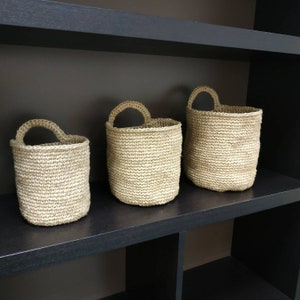 May include: Three woven baskets of varying sizes, made of natural fiber, with a light brown color, are displayed on a black shelf.