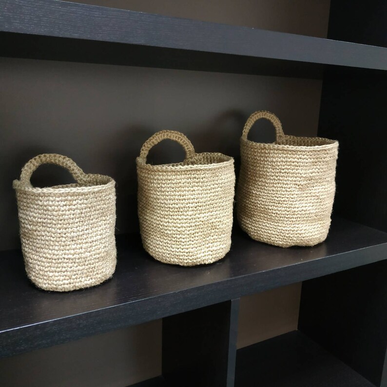 May include: Three woven baskets of varying sizes, made of natural fiber, with a light brown color, are displayed on a black shelf.