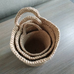 May include: Three nested, crocheted baskets made of natural jute fiber. The baskets have a natural beige color and are stacked inside each other. The largest basket has a handle.