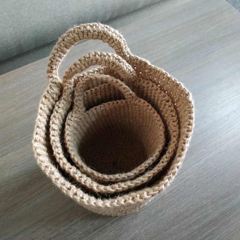 May include: Three nested, crocheted baskets made of natural jute fiber. The baskets have a natural beige color and are stacked inside each other. The largest basket has a handle.