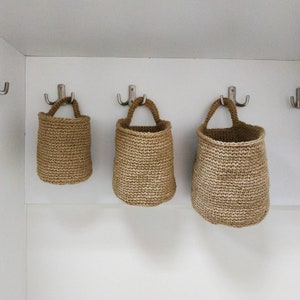 May include: Three brown crocheted hanging baskets with silver hooks on a white wall.