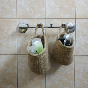 May include: Two beige crocheted hanging baskets with handles, filled with various toiletries, are attached to a chrome bar mounted on a tiled wall.