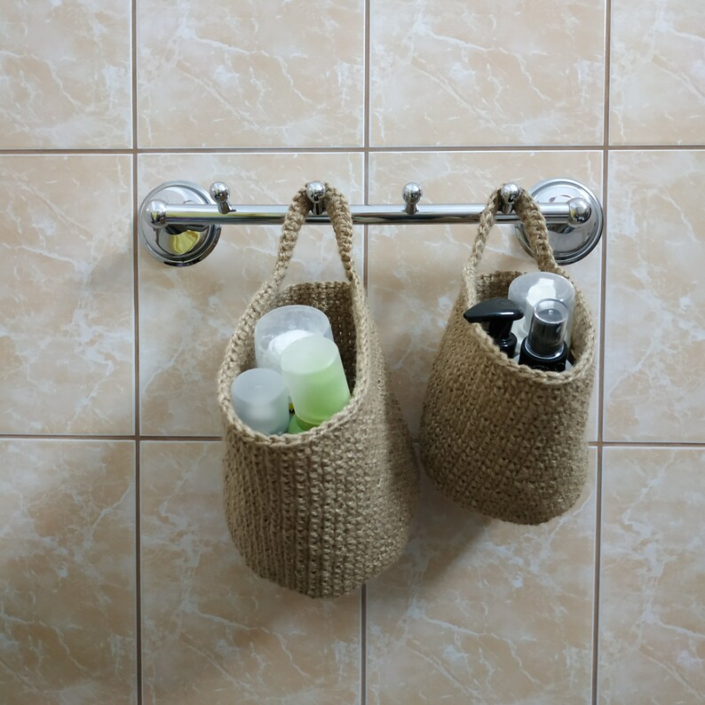 May include: Two beige crocheted hanging baskets with handles, filled with various toiletries, are attached to a chrome bar mounted on a tiled wall.
