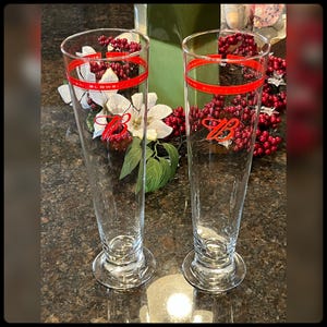 May include: Two tall, clear glass beer mugs with a red band around the top and a red letter "B" on the front. The mugs are sitting on a brown countertop.