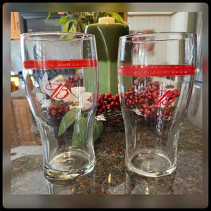 May include: Two clear glass beer mugs with a red band around the top and a red "Budweiser" logo. The mugs have a red "B" logo on the front and are sitting on a table with red berries and green leaves.