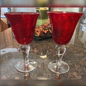 May include: Two red wine glasses with a bubble texture. The glasses have a flared bowl shape and sit on a clear, decorative stem and base. The glasses are made of glass and are on a dark countertop.