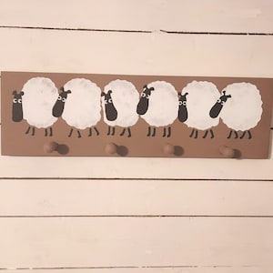 May include: A brown wooden wall-mounted coat rack featuring six cartoon sheep with white wool and black faces. The rack has six wooden pegs for hanging items. The sheep are evenly spaced across the rack.