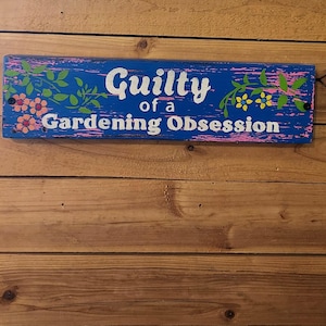 May include: A rectangular blue wooden sign with the words "Guilty of a Gardening Obsession" in white lettering. The sign is decorated with pink and yellow flowers and green leaves.