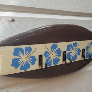 May include: A surfboard-shaped wall hook rack made of wood, featuring a dark brown finish on the top and bottom edges and a cream-colored center stripe. The center stripe is decorated with four blue hibiscus flower patterns and three black metal hooks.