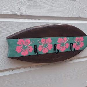 May include: A wooden surfboard-shaped key holder with a green and pink hibiscus flower design. The key holder has four black metal hooks.