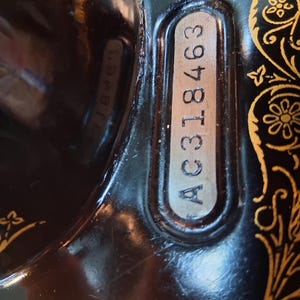 May include: Close-up of a vintage black sewing machine with gold floral detailing. The serial number AC318463 is visible on a silver plate. The machine's surface reflects the surrounding environment.