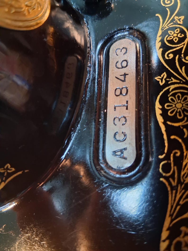 May include: Close-up of a vintage black sewing machine with gold floral detailing. The serial number AC318463 is visible on a silver plate. The machine's surface reflects the surrounding environment.