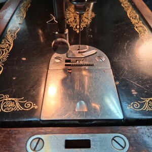 May include: Close-up of a vintage black sewing machine with gold floral accents. The needle is positioned over a silver plate, ready for stitching. The machine's wooden base features a metal plate with a screw.