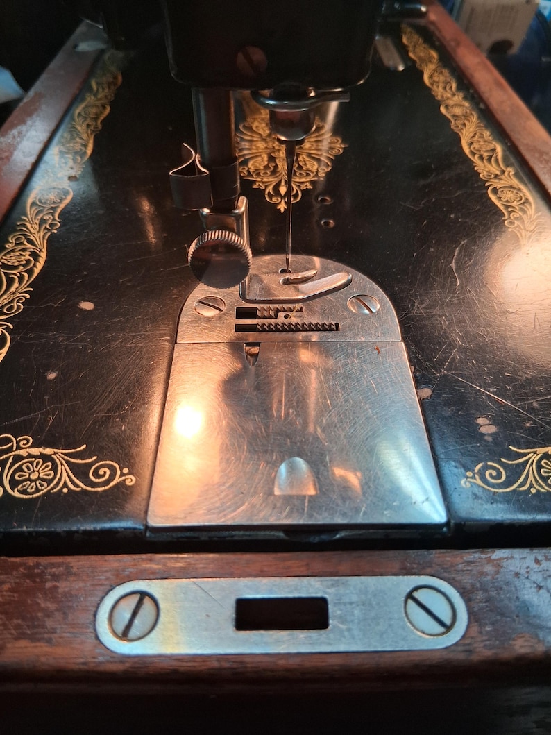 May include: Close-up of a vintage black sewing machine with gold floral accents. The needle is positioned over a silver plate, ready for stitching. The machine's wooden base features a metal plate with a screw.