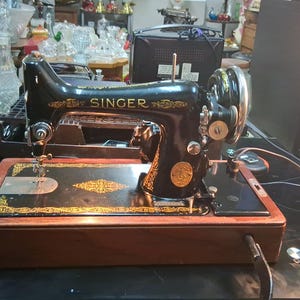 May include: A vintage Singer sewing machine, black with gold accents, sits on a wooden base. The machine features a decorative design and the Singer logo. The machine is on a black surface.