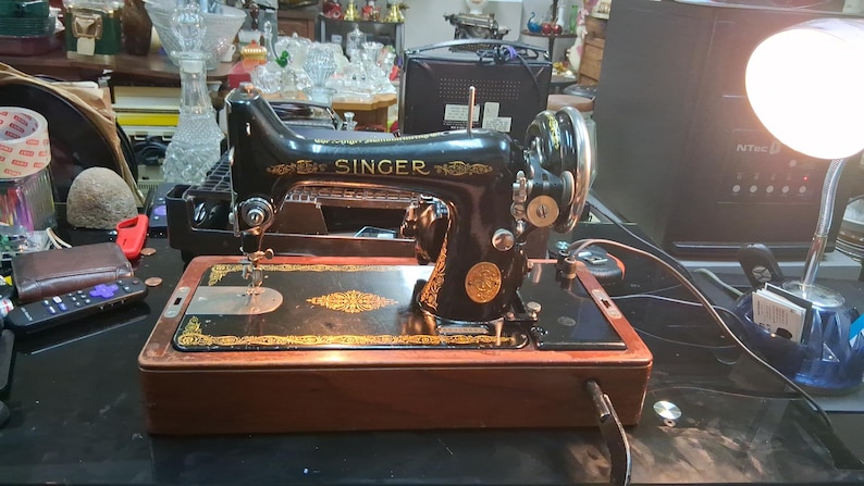 May include: A vintage Singer sewing machine, black with gold accents, sits on a wooden base. The machine features a decorative design and the Singer logo. The machine is on a black surface.