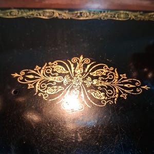 May include: Close-up of a vintage sewing machine with a black surface and gold filigree detailing. The gold design is centered and features floral and leaf motifs. The machine's surface shows signs of wear, adding to its antique charm.