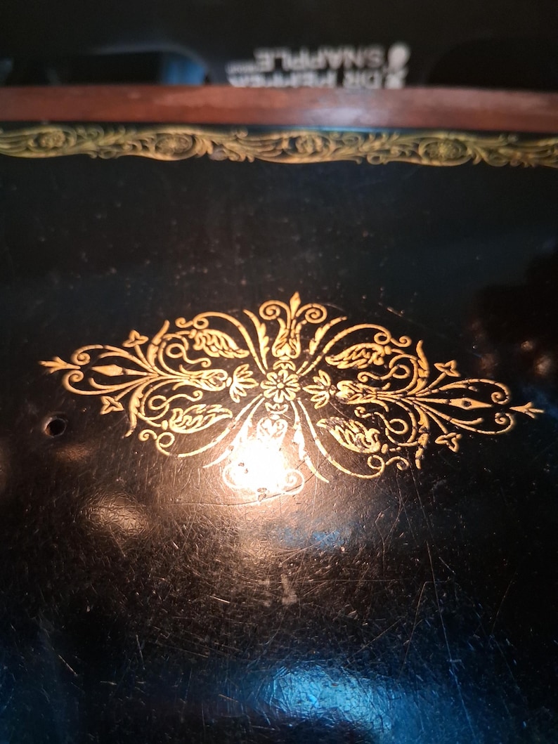 May include: Close-up of a vintage sewing machine with a black surface and gold filigree detailing. The gold design is centered and features floral and leaf motifs. The machine's surface shows signs of wear, adding to its antique charm.