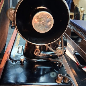 May include: An antique black sewing machine with a large, circular black component and a silver center. The machine is mounted on a wooden base with a shiny black surface. The machine has a belt and various mechanical parts.