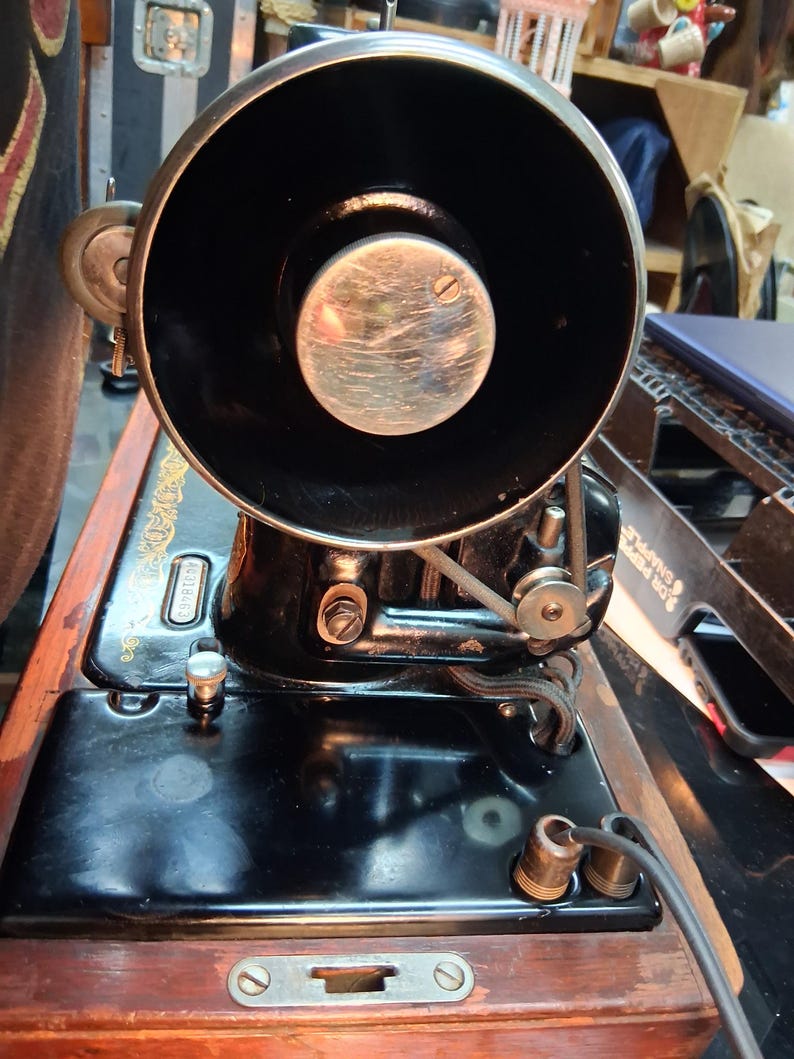 May include: An antique black sewing machine with a large, circular black component and a silver center. The machine is mounted on a wooden base with a shiny black surface. The machine has a belt and various mechanical parts.