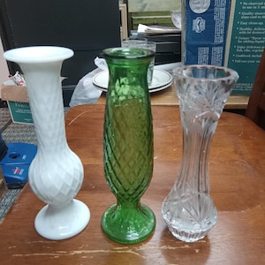 May include: Three decorative vases on a wooden table. The vases are white, green, and clear. The white vase has a bulbous shape with a textured pattern. The green vase is tall and slender with a diamond pattern. The clear vase has a floral design.