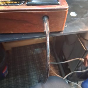 May include: Close-up of a vintage sewing machine with a wooden base and black metal components. The machine features a curved, metal foot pedal and a decorative emblem. The wooden base has a rich, brown color.