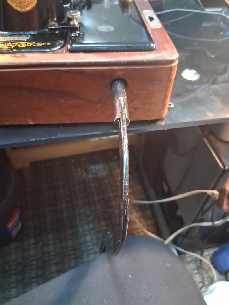 May include: Close-up of a vintage sewing machine with a wooden base and black metal components. The machine features a curved, metal foot pedal and a decorative emblem. The wooden base has a rich, brown color.