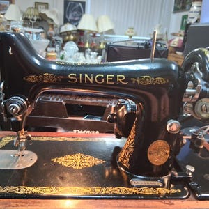 May include: A vintage Singer sewing machine, black with gold accents. The word "SINGER" is prominently displayed in gold lettering. The machine features intricate gold decorative patterns and a circular emblem.