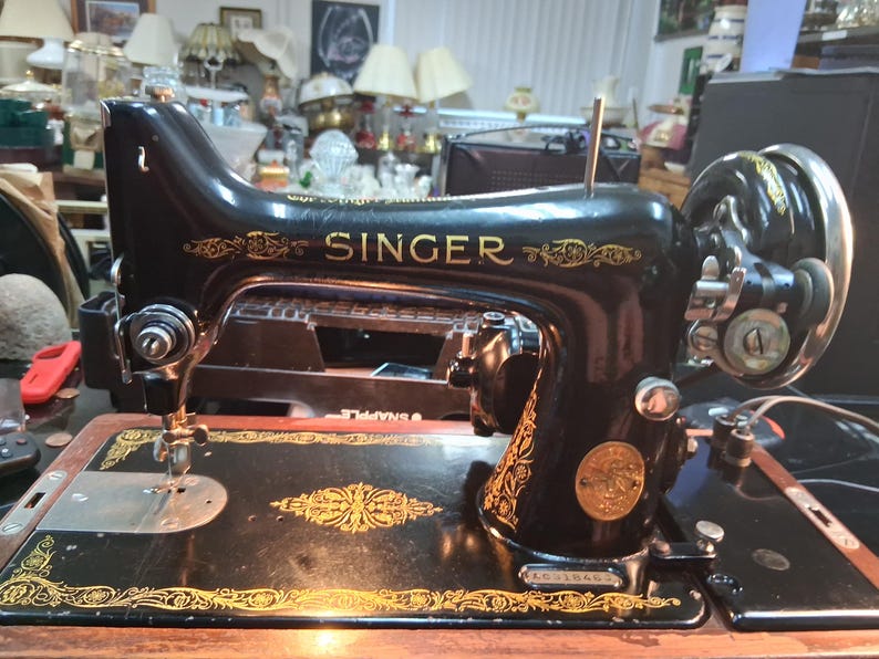 May include: A vintage Singer sewing machine, black with gold accents. The word "SINGER" is prominently displayed in gold lettering. The machine features intricate gold decorative patterns and a circular emblem.