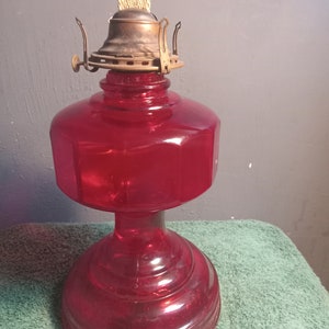 May include: A red glass oil lamp with a brass burner and a wick. The lamp has a round base and a cylindrical body.