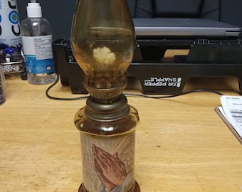 Puede incluir: Una lámpara de aceite vintage con una chimenea de vidrio transparente y una base de madera. La base presenta una imagen impresa de dos manos juntas en oración.