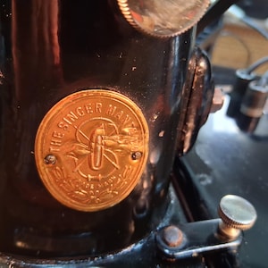 May include: Close-up of a vintage black Singer sewing machine. A gold-colored emblem with the text "THE SINGER MFG. CO." is affixed to the machine's body. The emblem features a stylized design and is secured with two small screws.
