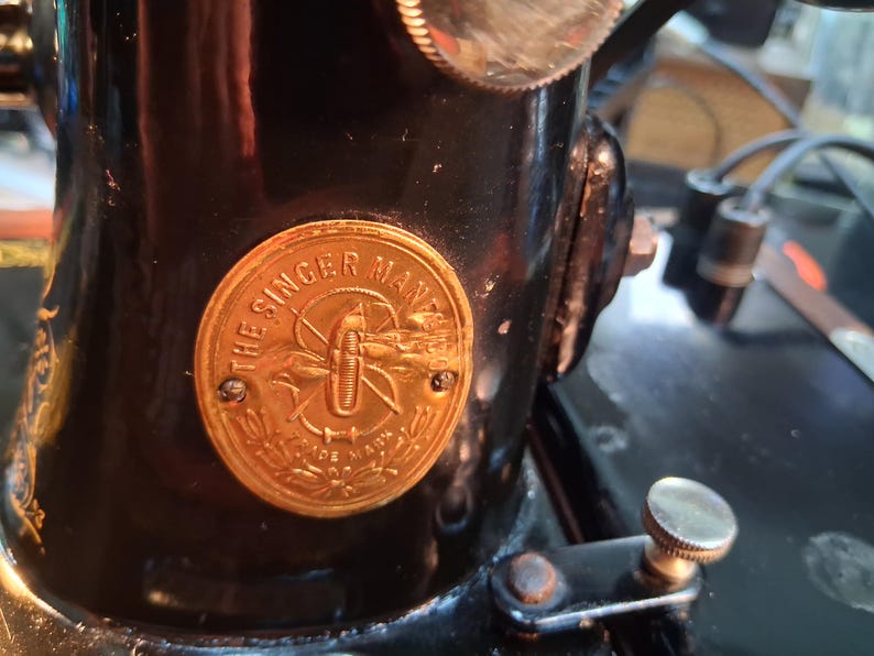 May include: Close-up of a vintage black Singer sewing machine. A gold-colored emblem with the text "THE SINGER MFG. CO." is affixed to the machine's body. The emblem features a stylized design and is secured with two small screws.
