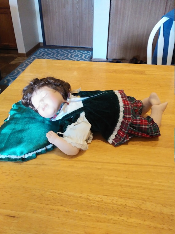 Porcelain Doll #118 baby laying down on pillow wearing green velvet top and a red and green flannel bottom. Circa. 1980s