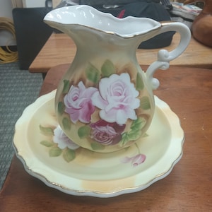 May include: A white porcelain pitcher with a gold trim and a floral design featuring pink roses. The pitcher is sitting on a matching saucer with a scalloped edge.