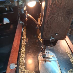 May include: Close-up of a vintage Singer sewing machine. The black machine features gold floral detailing and a decorative, ornate design on the head. A small lamp is attached, illuminating the needle and work area.