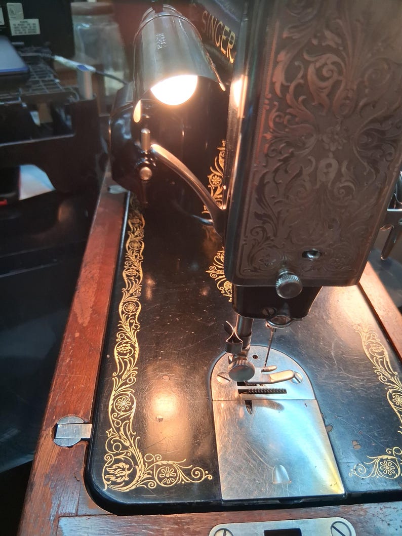 May include: Close-up of a vintage Singer sewing machine. The black machine features gold floral detailing and a decorative, ornate design on the head. A small lamp is attached, illuminating the needle and work area.