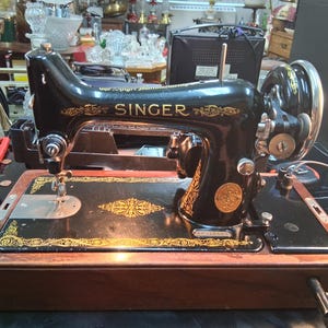 May include: Black Singer sewing machine with gold accents. The machine features the Singer logo and decorative patterns. The base is a dark wood color. The machine is on a table with other items in the background.