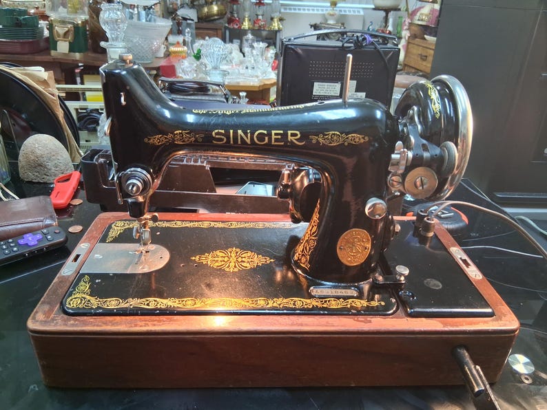 May include: Black Singer sewing machine with gold accents. The machine features the Singer logo and decorative patterns. The base is a dark wood color. The machine is on a table with other items in the background.