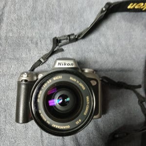May include: A silver Nikon camera with a black lens and a black strap. The lens is a 28-90mm 1:3.5-5.6 macro lens. The camera is made in Japan.