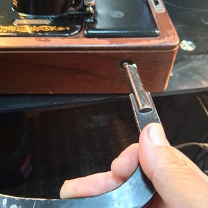 May include: Close-up of a vintage black Singer sewing machine with gold accents. The wooden base shows signs of wear. A black metal lever is being inserted into the machine's side. The machine's design is classic and ornate.