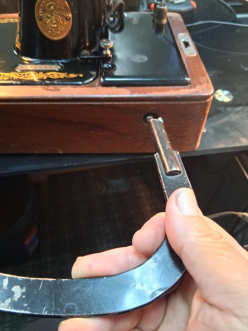 May include: Close-up of a vintage black Singer sewing machine with gold accents. The wooden base shows signs of wear. A black metal lever is being inserted into the machine's side. The machine's design is classic and ornate.