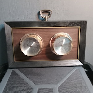 Puede incluir: Un reloj de pared de madera enmarcado con dos indicadores analógicos redondos. Los indicadores tienen biseles dorados y manecillas negras. El indicador izquierdo está etiquetado como "Humedad" y el indicador derecho está etiquetado como "Termómetro".