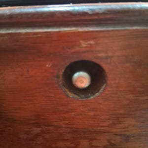 May include: Close-up of a section of a wooden object, possibly furniture. The wood has a rich, reddish-brown color with visible grain. A circular hole reveals a metal component, likely a screw or bolt, adding a contrasting metallic element.