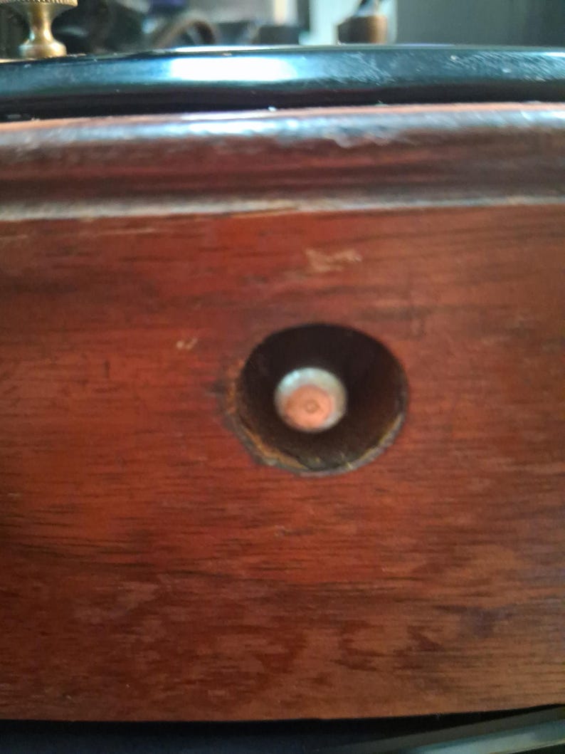 May include: Close-up of a section of a wooden object, possibly furniture. The wood has a rich, reddish-brown color with visible grain. A circular hole reveals a metal component, likely a screw or bolt, adding a contrasting metallic element.