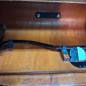 May include: An antique black metal and wood tool, possibly for woodworking, resting inside a wooden cabinet. The tool has a curved arm and a blade-like component. The cabinet is made of light brown wood.
