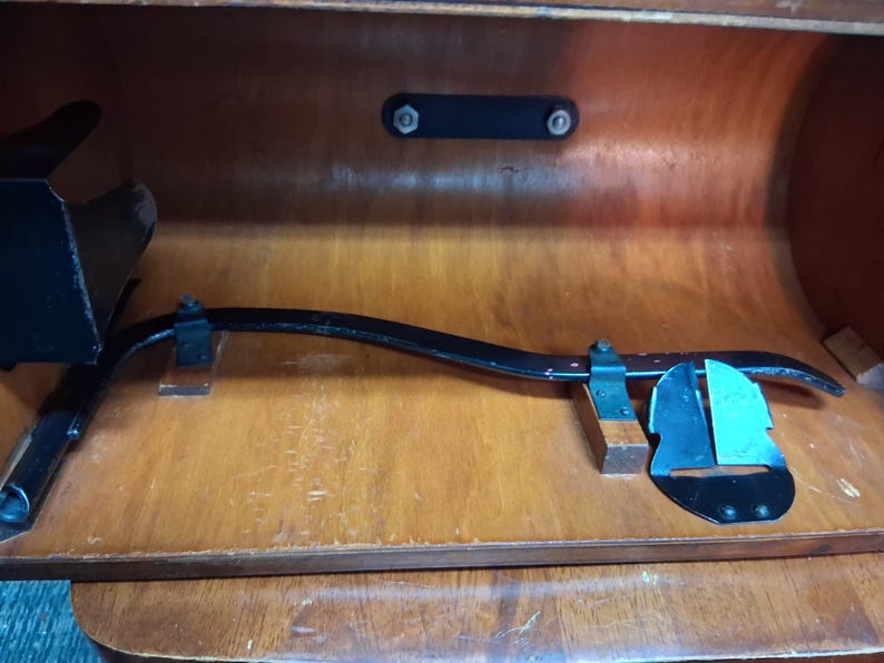 May include: An antique black metal and wood tool, possibly for woodworking, resting inside a wooden cabinet. The tool has a curved arm and a blade-like component. The cabinet is made of light brown wood.