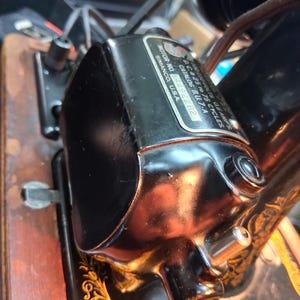 May include: Close-up of a black sewing machine motor with a shiny, reflective surface. The motor has a label with text including "SIMANCO, U.S.A." and "CATALOG BU.7-A". The motor is attached to a wooden base with gold decorative accents.