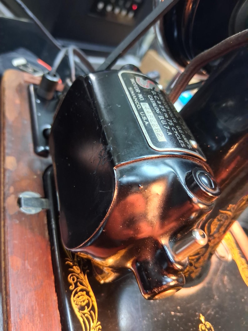 May include: Close-up of a black sewing machine motor with a shiny, reflective surface. The motor has a label with text including "SIMANCO, U.S.A." and "CATALOG BU.7-A". The motor is attached to a wooden base with gold decorative accents.